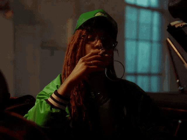 a woman with dreadlocks wearing a green jacket and hat smoking a cigarette