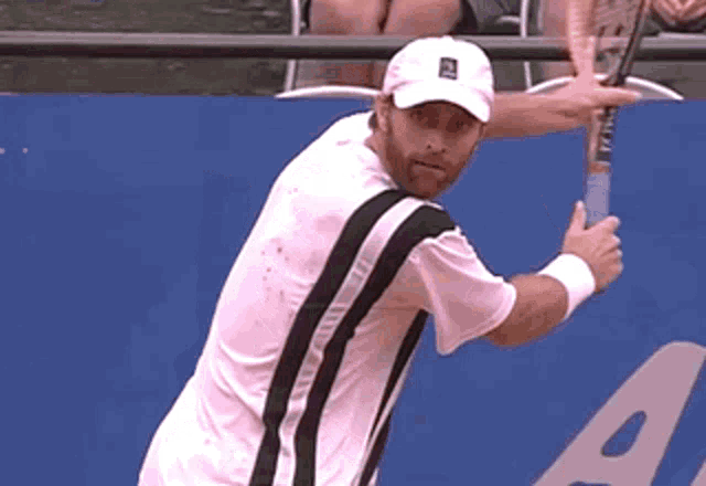 a man in a white hat holds a tennis racquet in front of a blue wall with the letter a on it