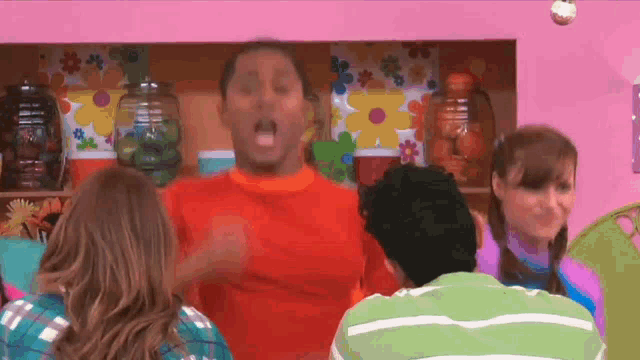 a man in an orange shirt is standing in front of a pink wall with flowers on it