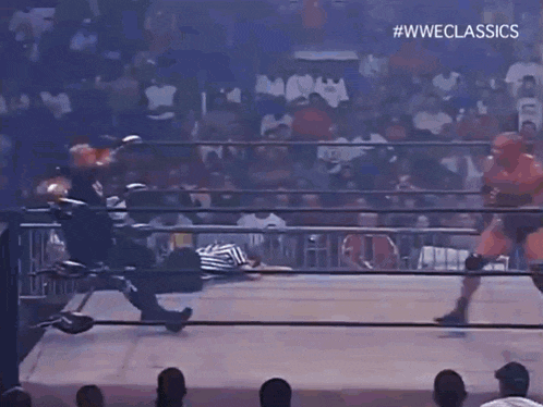 a man in a black shirt with the letter b on it is standing next to a wrestler in a ring .