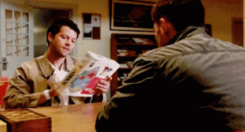 two men are sitting at a table talking to each other while one of them is reading a magazine .