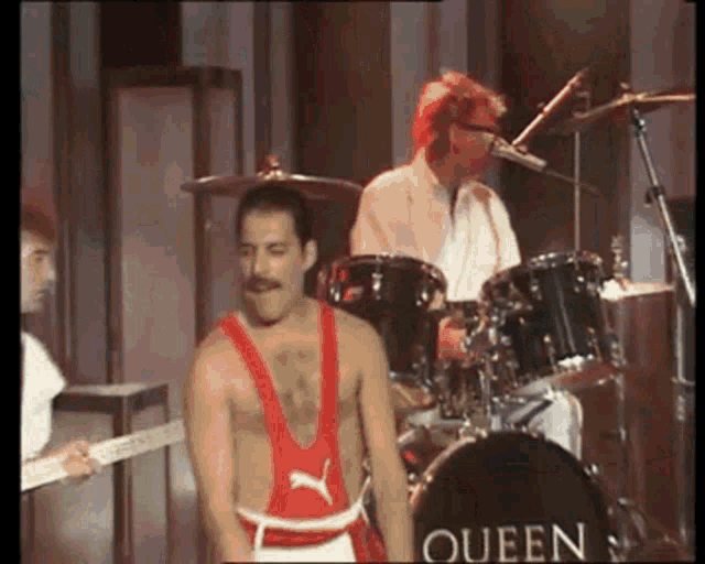 a man in a red tank top is standing in front of a drum set in a queen band .