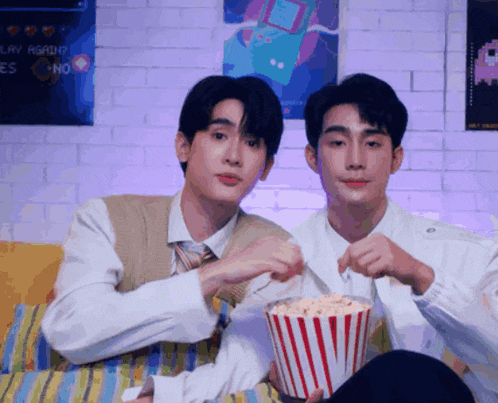 two men sitting on a couch eating popcorn with a poster that says " play again " on the wall behind them
