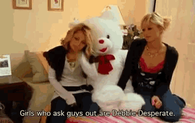two women sitting next to a teddy bear with the words girls who ask guys out are debbie desperate on the bottom