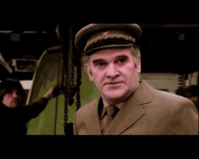 a man in a suit and hat looks at the camera with another man in the background