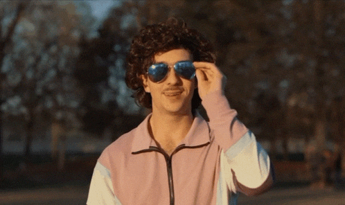 a man wearing sunglasses and a pink jacket looks at the camera