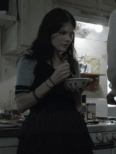 a woman is eating a bowl of cereal in a kitchen with a box of honey bunches of oats on the stove