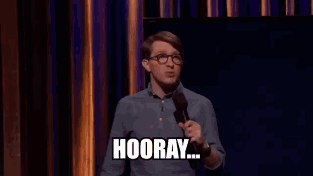 a man in glasses is holding a microphone and saying `` hooray '' .