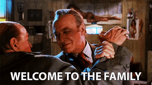a man in a suit and tie is hugging another man in a room with the words welcome to the family .