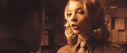 a woman in a brown shirt and tie looks at a man in a dark room