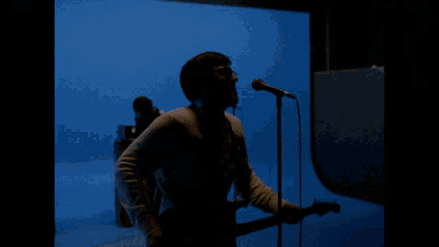 a man playing a guitar and singing into a microphone in front of a blue wall