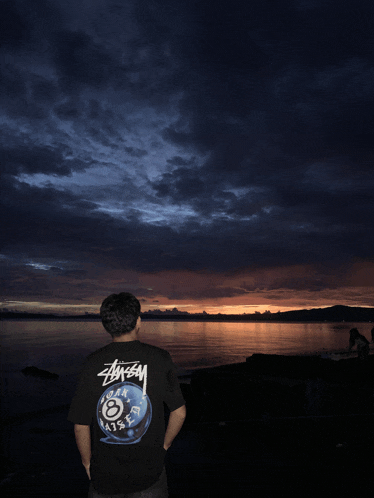 a man wearing a stussy shirt stands in front of a body of water