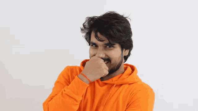 a man with a beard is wearing an orange hoodie and covering his mouth with his hand