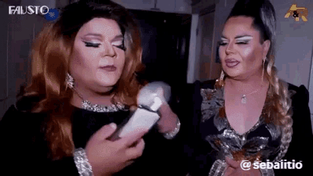 two drag queens are standing next to each other and looking at a cell phone .