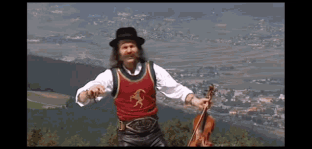 a man in a hat is holding a violin in front of a mountain range