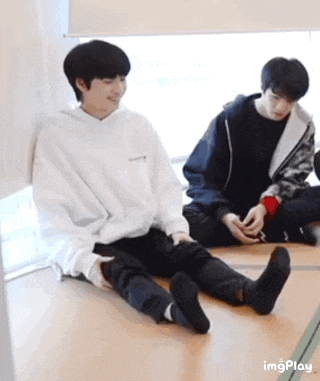 two young men are sitting on the floor with their legs crossed and one is holding a cell phone .