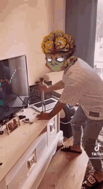 a man with a mask on his head is standing in front of a laptop and a television .