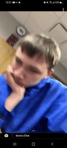 a man in a blue shirt covering his mouth with his hand and a clock on the wall behind him