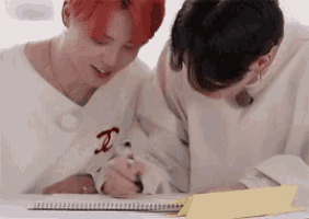 two young men are sitting at a table writing in a notebook with a pen