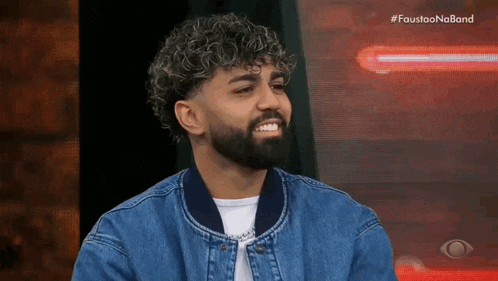 a man with curly hair and a beard wearing a denim jacket is smiling .