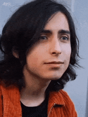 a close up of a young man with long hair wearing an orange jacket and a black shirt .