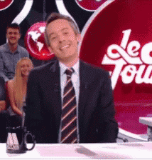 a man in a suit and tie is smiling in front of a red sign that says le tour
