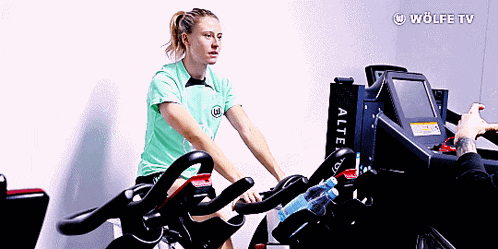 a woman in a green shirt is riding an exercise bike with a wolfe tv logo in the background