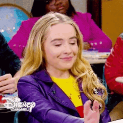a girl wearing a purple jacket and a yellow t-shirt is sitting in a disney channel classroom