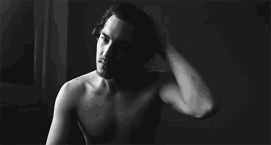 a black and white photo of a shirtless man touching his hair in a room .