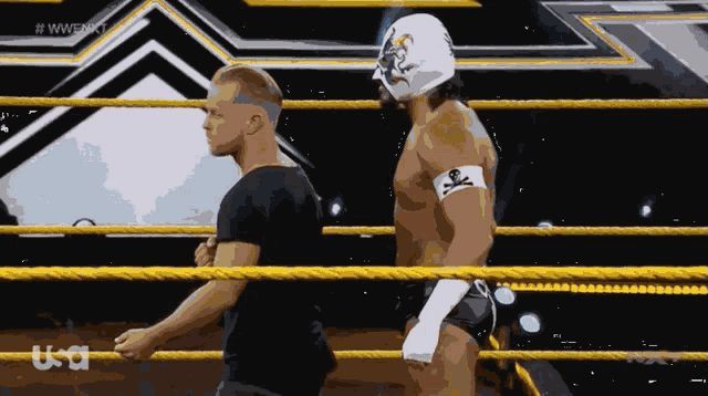 a wrestler with a skull and crossbones tattoo on his arm talks to another wrestler in a wrestling ring