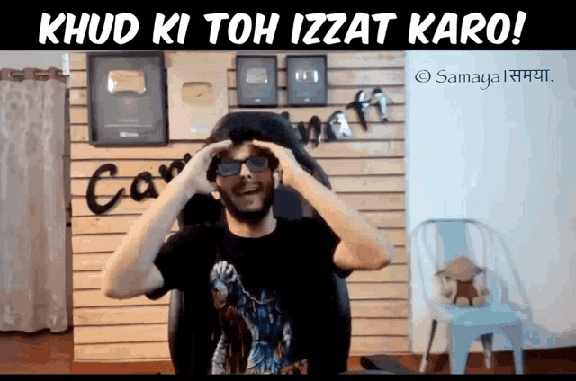 a man wearing sunglasses and a black shirt is sitting in front of a wall that says ' samaya '