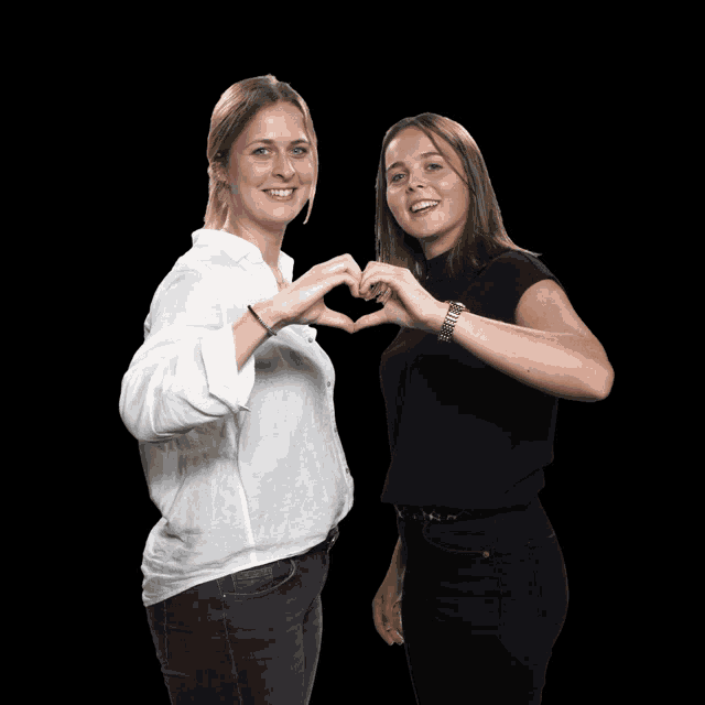two women make a heart shape with their hands against a black background