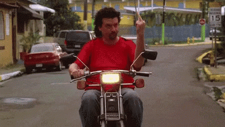 a man in a red shirt is riding a motorcycle down a street giving the middle finger .