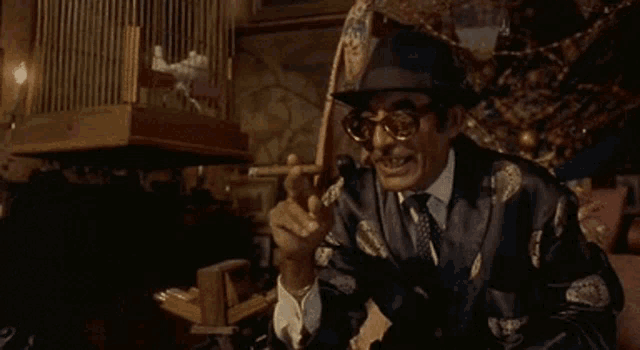a man in a hat and sunglasses is smoking a cigar and pointing at the camera .