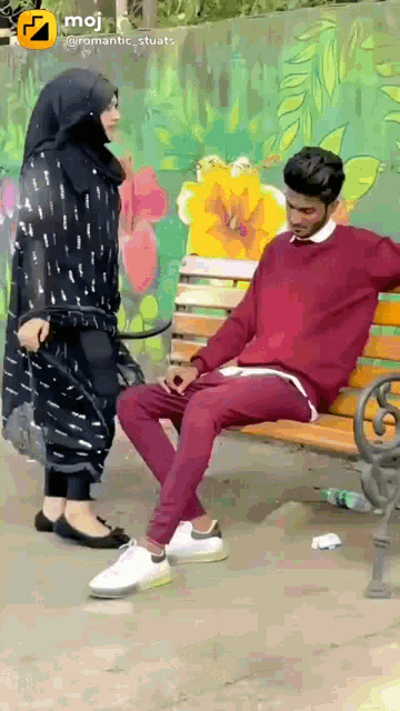 a woman in a hijab stands next to a man sitting on a bench in front of a wall with flowers on it