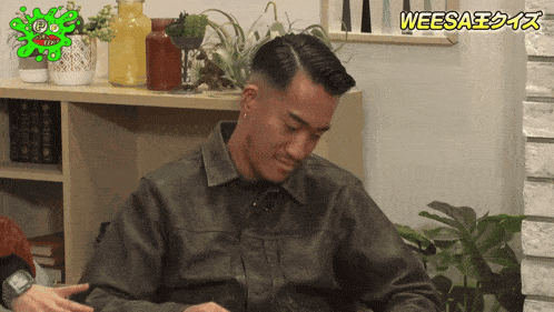 a man in a leather jacket is sitting in front of a shelf with plants and a green sticker that says weesa