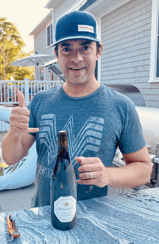a man wearing a blue hat holds up a bottle of benovia wine
