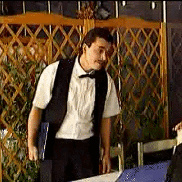 a man in a tuxedo and bow tie is standing in front of a table holding a menu .