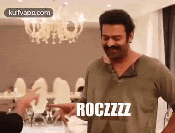 a man with a mustache is standing in a room with a chandelier in the background and making a funny face .
