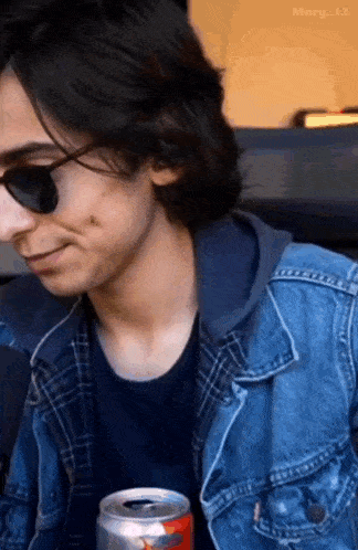 a young man wearing sunglasses and a denim jacket is holding a soda can .