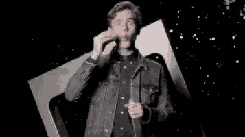 a man in a denim jacket is standing in front of a black background and holding a glass of wine .