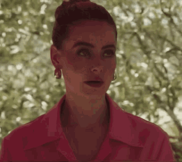 a woman in a pink shirt and earrings is standing in front of trees .