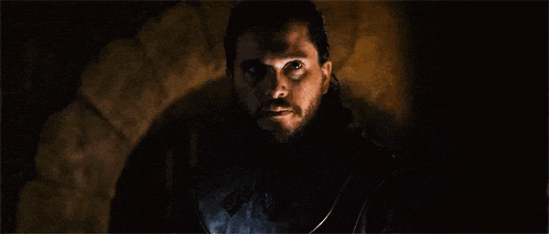 a man with a beard and long hair is standing in front of a fireplace in a dark room .