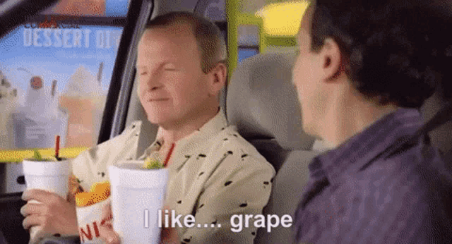 two men are sitting in a car and one of them is holding a cup that says grape