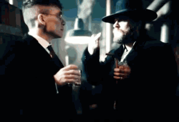 a man in a suit and tie is holding a glass of whiskey while talking to another man in a hat .