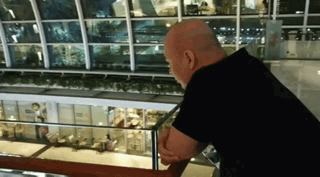 a bald man in a black shirt is standing on a balcony overlooking a shopping mall