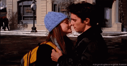 a man and a woman are kissing in front of a building . the woman is wearing a blue hat .