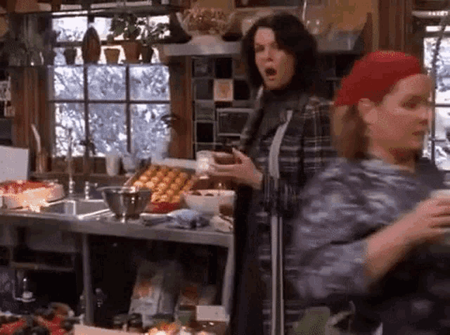 two women are standing in a kitchen talking to each other . one woman is holding a cup of coffee .