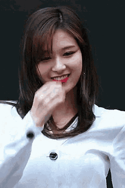 a woman wearing a white shirt and red lipstick is smiling and covering her mouth with her hand .