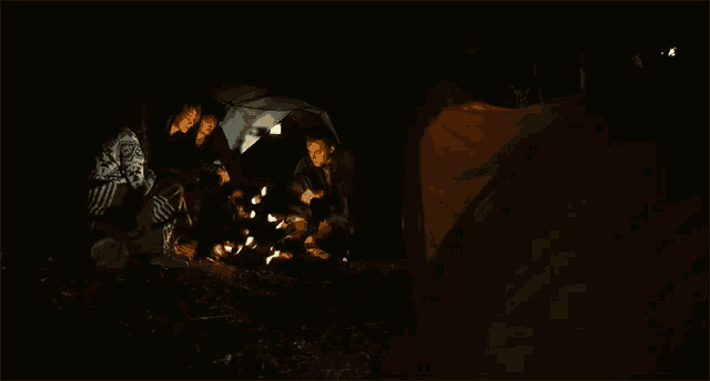 a group of people are sitting around a campfire with a tent in the background